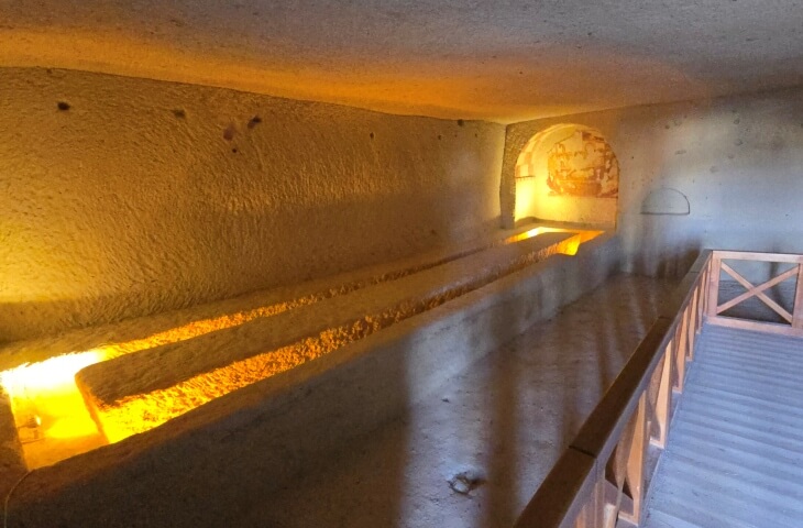 Çarıklı Kilise’nin yemekhanesi. Kaya oyma uzun masa ve oturma alanları, ışıklandırılmış şekilde görülüyor. Arka duvarda silik fresk izleri mevcut.
